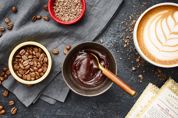 3 bowls on a table filled with chocolate ganache, coffee beans, hot chocolate mix. A mocha with flower latte art on the right.
