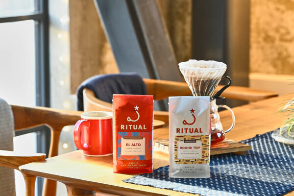 Two 12 ounce bags of coffee with a ceramic red mug and glass v60 brewing coffee